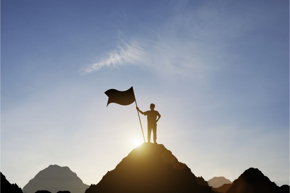 Silhouette of businessman holding flag on the top of mountain with over blue sky and sunlight